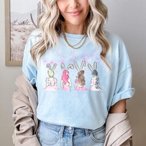 Puede incluir: Una camiseta azul claro con un gr&aacute;fico de cuatro mujeres con orejas de conejo y vestidos rosas. El gr&aacute;fico es una ilustraci&oacute;n de estilo acuarela sobre un fondo azul claro.