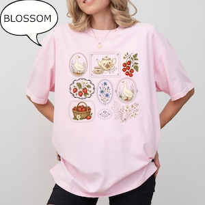 Puede incluir: Una camiseta rosa claro con un estampado gr&aacute;fico de varias ilustraciones enmarcadas que incluyen gansos, una tetera, fresas en una cesta, tomates en rama y ramitas de flores. La palabra BLOSSOM aparece en un globo de texto sobre la camiseta.