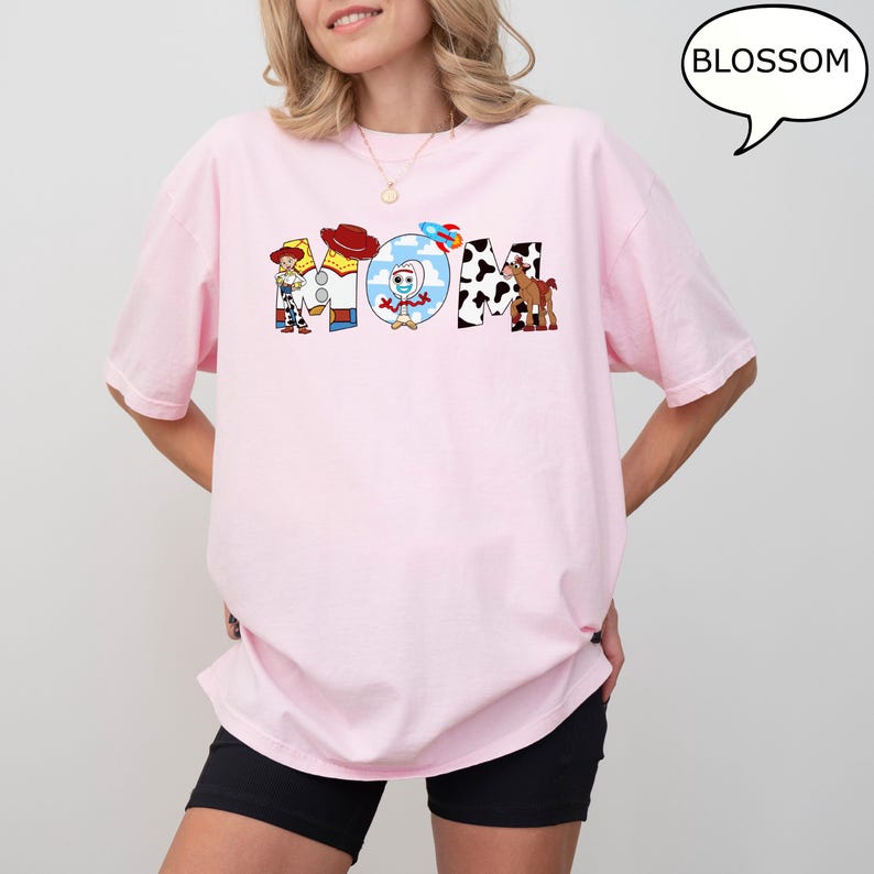 May include: A pale pink t-shirt with the word "MOM" in a colourful cartoon design. The "M" features a cowboy, the "O" has a character in a circle, and the "M" has a cow print. The shirt has short sleeves.