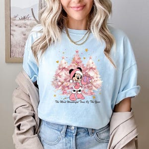 May include: Light blue t-shirt featuring a Christmas-themed graphic. The design includes a cartoon character in a Santa hat holding a gift, with pink Christmas trees and the text "The Most Wonderful Time Of The Year".