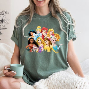 Puede incluir: Una camiseta verde con un estampado gr&aacute;fico de princesas Disney, incluyendo Cenicienta, Ariel, Bella y Moana. Las princesas est&aacute;n todas sonriendo y mirando al espectador.