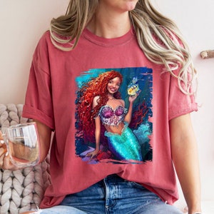 May include: A red t-shirt featuring a graphic of a mermaid with long red hair holding a yellow fish. The mermaid is wearing a pink and purple seashell bra and a turquoise tail.