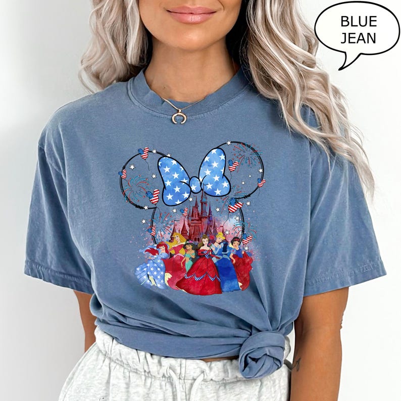 Puede incluir: Camiseta azul vaquero con un dise&ntilde;o de Minnie Mouse con un lazo patri&oacute;tico y fuegos artificiales. El dise&ntilde;o incluye un castillo y varias princesas de Disney. El texto "BLUE JEAN" est&aacute; en una burbuja.