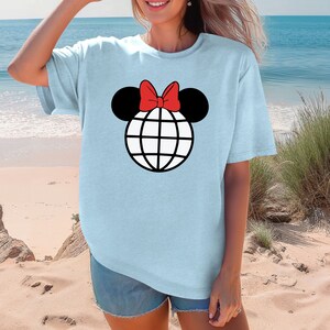 K&ouml;nnte beinhalten: Hellblaues T-Shirt mit einem schwarzen und wei&szlig;en Globus-Motiv, das mit einer roten Schleife verziert ist und an Mickey Mouse-Ohren erinnert.