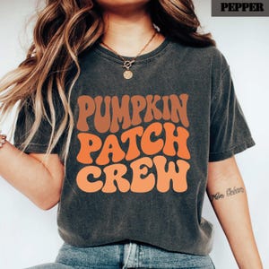 May include: A charcoal gray short-sleeve t-shirt featuring the text PUMPKIN PATCH CREW in a wavy, retro-style font. The lettering is displayed in shades of orange and brown. The shirt has a crew neckline and a vintage-washed appearance.