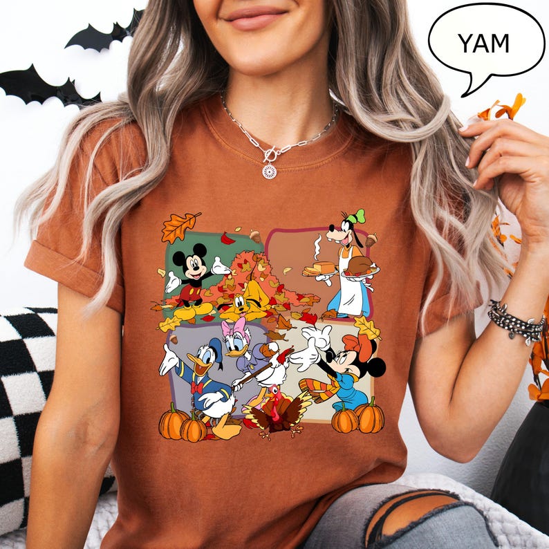 May include: A rust-colored t-shirt featuring a Thanksgiving-themed graphic with cartoon characters. The design includes Mickey Mouse, Minnie Mouse, Donald Duck, Daisy Duck, and Goofy, along with pumpkins and autumn leaves. The word "YAM" is in a speech bubble.