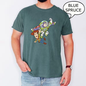 May include: A blue spruce t-shirt featuring a graphic of Buzz Lightyear and Woody from Toy Story. The characters are in mid-air, with Buzz Lightyear in a flying pose. The text "BLUE SPRUCE" is in a speech bubble.