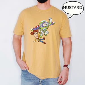 May include: Mustard-colored t-shirt featuring a graphic of Woody and Buzz Lightyear from Toy Story. The cartoon characters are depicted in mid-air, with Buzz Lightyear's wings extended. The word "MUSTARD" is in a speech bubble.