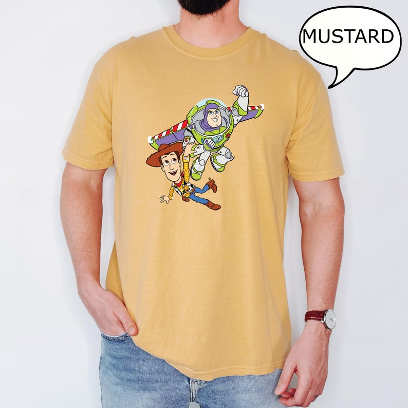 May include: Mustard-colored t-shirt featuring a graphic of Woody and Buzz Lightyear from Toy Story. The cartoon characters are depicted in mid-air, with Buzz Lightyear's wings extended. The word "MUSTARD" is in a speech bubble.