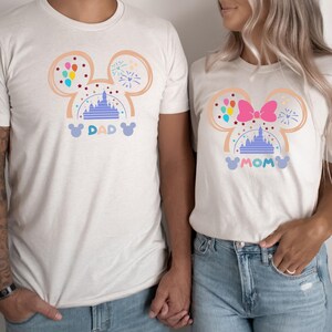 Pode incluir: Camisas brancas com um design de silhueta do Mickey Mouse que apresenta um castelo roxo, bal&otilde;es e fogos de artif&iacute;cio. As camisas dizem "DAD" e "MOM" em uma fonte caprichosa.