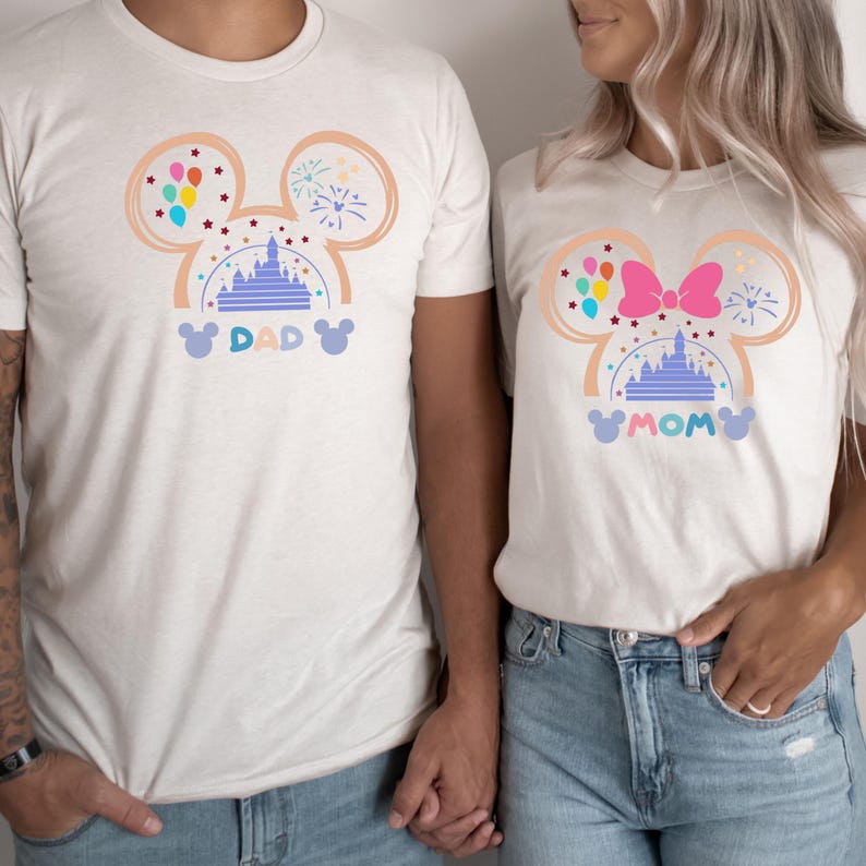 Pode incluir: Camisas brancas com um design de silhueta do Mickey Mouse que apresenta um castelo roxo, bal&otilde;es e fogos de artif&iacute;cio. As camisas dizem "DAD" e "MOM" em uma fonte caprichosa.
