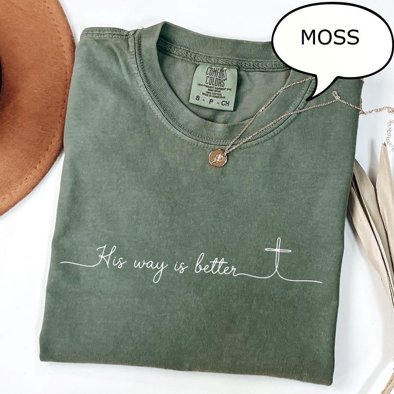 May include: A moss green t-shirt with the text "His way is better" in white script and a cross graphic. The shirt has a crew neck and short sleeves. A silver necklace with a pendant is draped across the shirt.
