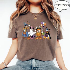 May include: Espresso-colored t-shirt featuring a Halloween-themed graphic of Disney characters in costumes. Goofy, Minnie Mouse, Mickey Mouse, Donald Duck, and other characters are depicted with pumpkins and ghosts. The word "ESPRESSO" is in a speech bubble.