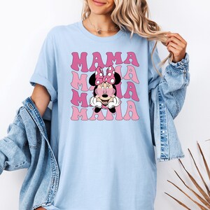 Puede incluir: Camiseta azul claro con un gr&aacute;fico rosa de Minnie Mouse con gafas de sol en forma de coraz&oacute;n y el texto "MAMA" repetido en una fuente retro.
