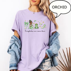 K&ouml;nnte beinhalten: Ein helllila T-Shirt mit einer gr&uuml;nen und goldenen Grafik einer Prinzessin, eines Frosches und eines Schuhs. Der Text "Fairytales can come true" steht darunter. Das Wort "ORCHID" steht in einer Sprechblase.