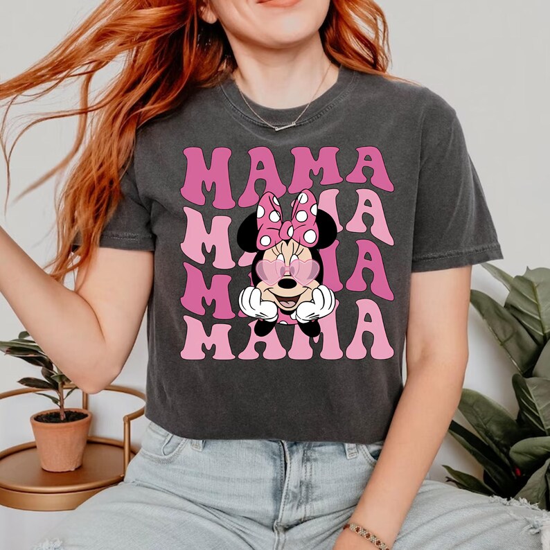 Puede incluir: Una camiseta negra recortada con un gr&aacute;fico rosa de Minnie Mouse con gafas de sol en forma de coraz&oacute;n y el texto "MAMA" repetido en un estilo retro.