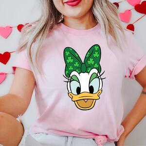 Puede incluir: Camiseta rosa claro con un dise&ntilde;o de dibujos animados de Daisy Duck con un lazo verde adornado con tr&eacute;boles. La camiseta tiene mangas cortas y cuello redondo. El dise&ntilde;o est&aacute; centrado en la parte delantera.