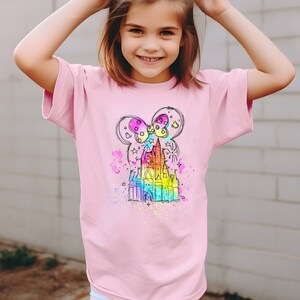 Puede incluir: Una camiseta rosa con un boceto de un castillo de color arco&iacute;ris con un lazo en la parte superior. El castillo est&aacute; rodeado de estrellas y fuegos artificiales.