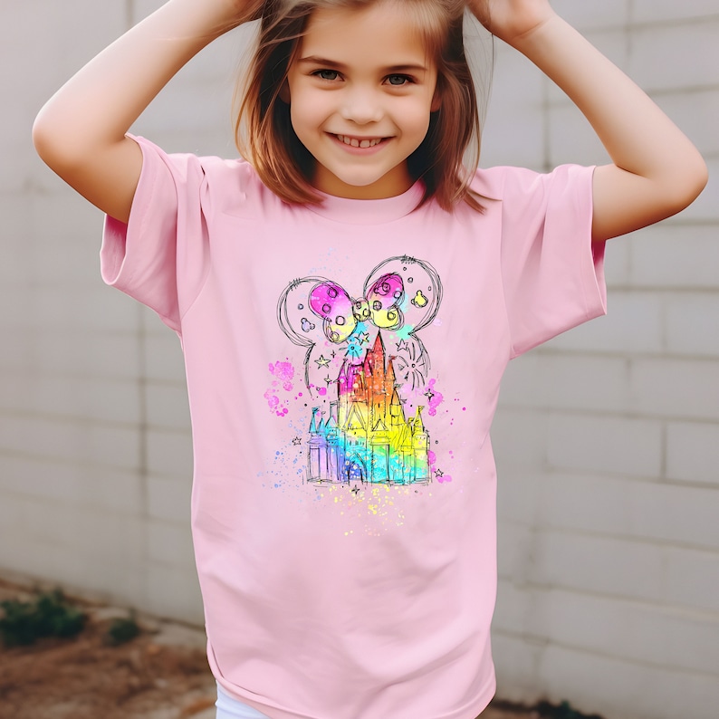 Puede incluir: Una camiseta rosa con un boceto de un castillo de color arco&iacute;ris con un lazo en la parte superior. El castillo est&aacute; rodeado de estrellas y fuegos artificiales.