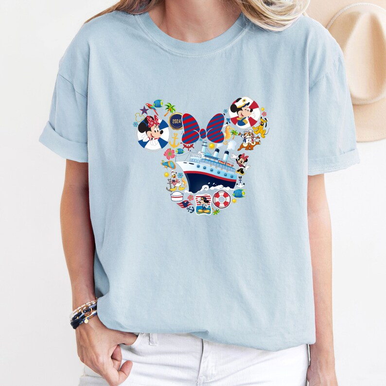 Pu&ograve; includere: T-shirt azzurra con un design a forma di testa di Topolino riempito di elementi nautici. Il design include una nave da crociera, Minnie Mouse e altri personaggi. L'anno "2024" &egrave; incluso.