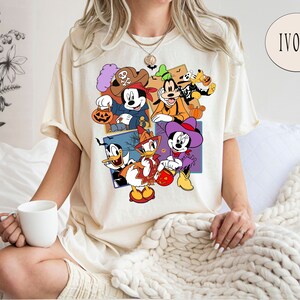 K&ouml;nnte beinhalten: Ein cremefarbenes T-Shirt mit einem grafischen Druck von Mickey Mouse, Minnie Mouse, Donald Duck, Goofy und Pluto in Halloween-Kost&uuml;men. Der Text "IVORY" ist in der oberen rechten Ecke des Bildes sichtbar.