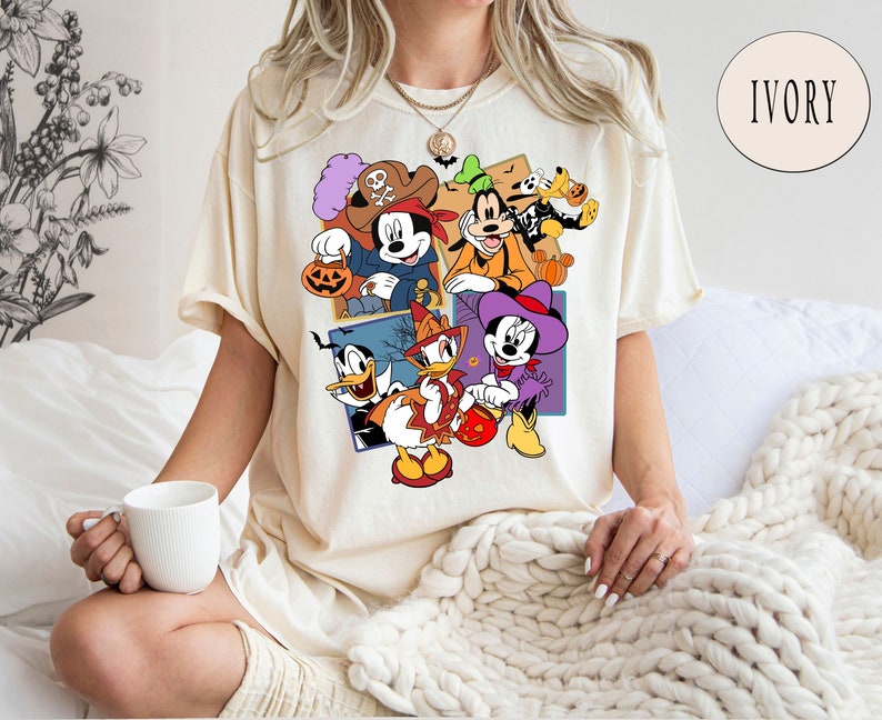 K&ouml;nnte beinhalten: Ein cremefarbenes T-Shirt mit einem grafischen Druck von Mickey Mouse, Minnie Mouse, Donald Duck, Goofy und Pluto in Halloween-Kost&uuml;men. Der Text "IVORY" ist in der oberen rechten Ecke des Bildes sichtbar.