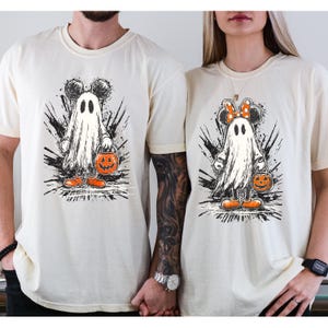 May include: Two cream-colored t-shirts featuring a Halloween ghost design. The ghost has Mickey Mouse ears and is holding a jack-o'-lantern bucket. The design is in black, white, and orange. The shirts are short-sleeved.