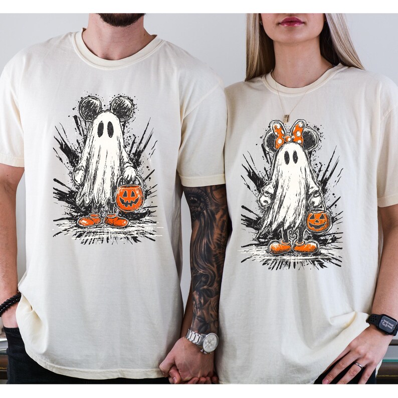 May include: Two cream-colored t-shirts featuring a Halloween ghost design. The ghost has Mickey Mouse ears and is holding a jack-o'-lantern bucket. The design is in black, white, and orange. The shirts are short-sleeved.