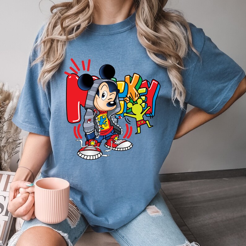 Pode incluir: Uma camiseta azul com um gr&aacute;fico de Mickey Mouse vestindo uma jaqueta vermelha e azul, cal&ccedil;as pretas e t&ecirc;nis vermelhos e brancos. O gr&aacute;fico tamb&eacute;m inclui a palavra "Mickey" em letras coloridas.