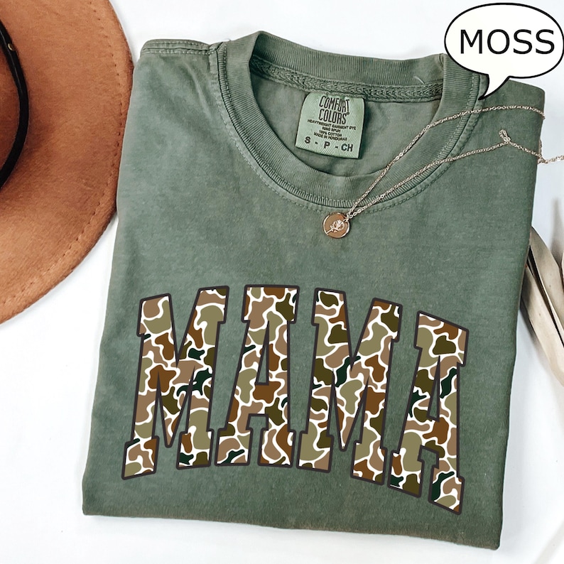 May include: A green t-shirt with the word "MAMA" in a camouflage pattern. The shirt is made of a soft, comfortable material and has a relaxed fit.