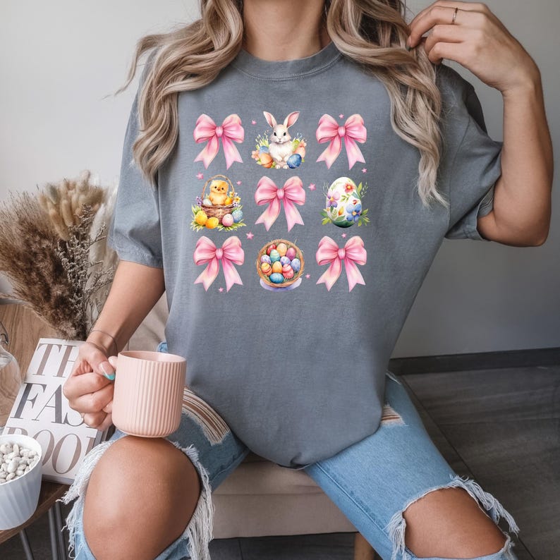 Puede incluir: Una camiseta gris con un dise&ntilde;o gr&aacute;fico de lazos rosas, huevos de Pascua, un conejito y un pollito. El dise&ntilde;o se repite en un patr&oacute;n de cuadr&iacute;cula.