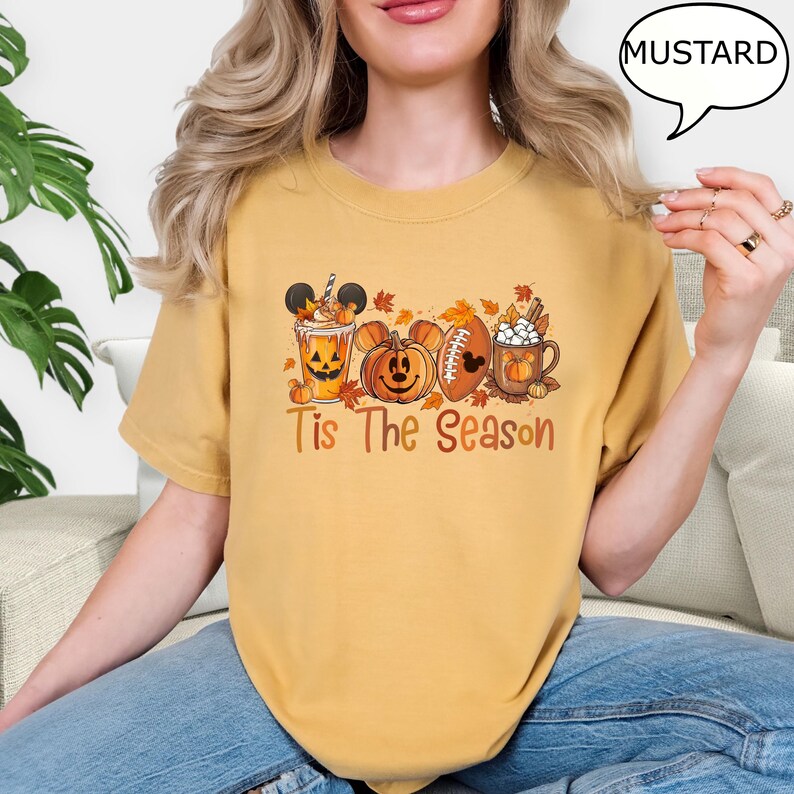May include: Mustard-colored t-shirt with a fall-themed graphic. The design includes a pumpkin, a football, and a mug of hot chocolate, all with Mickey Mouse ears. The text "Tis The Season" is printed below the graphic.