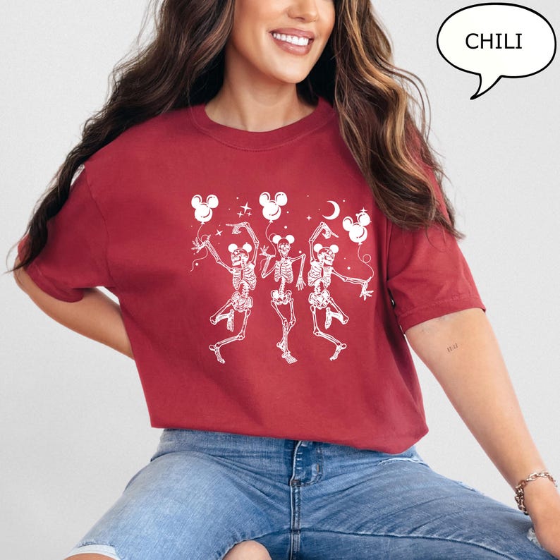 May include: A maroon t-shirt featuring a white graphic of three dancing skeletons holding Mickey Mouse-shaped balloons. The word "CHILI" is in a speech bubble. The shirt is made of a soft material.