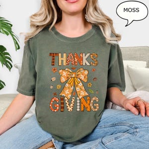 May include: A moss green t-shirt with the words "THANKS GIVING" in a fall-themed design. The design includes a large bow, flowers, and fall leaves. The shirt is a comfortable, casual style.