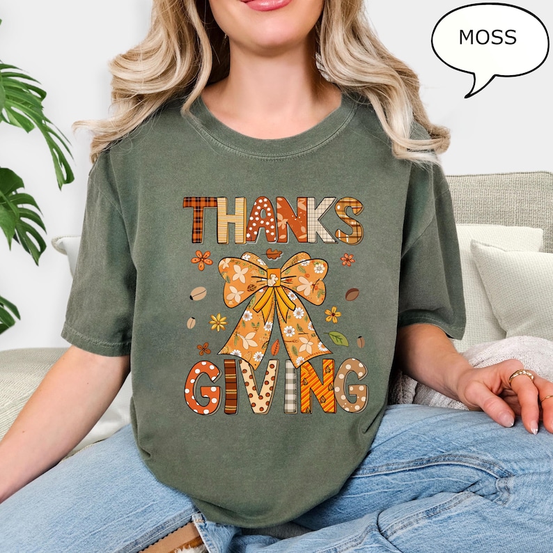 May include: A moss green t-shirt with the words "THANKS GIVING" in a fall-themed design. The design includes a large bow, flowers, and fall leaves. The shirt is a comfortable, casual style.