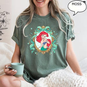 May include: Moss green t-shirt featuring a framed illustration of Ariel from The Little Mermaid. The illustration shows Ariel with red hair, a blue top, and a green tail. The frame is gold with teal ribbons and accents. The word "MOSS" is in a speech bubble.