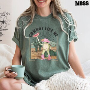 May include: A green t-shirt with the text "COWBOY LIKE ME" in a vintage style font. A cartoon frog wearing a pink cowboy hat and boots is holding a watermelon slice. The frog is standing in a desert landscape with cacti.