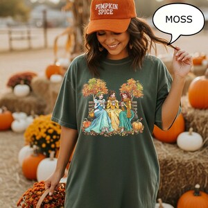 May include: Dark green t-shirt with a graphic of three princesses sitting on a bench under autumn trees. The princesses are wearing colorful gowns and holding mugs. The image includes pumpkins and fall foliage. The person in the image is wearing an orange cap that says "Pumpkin Spice".