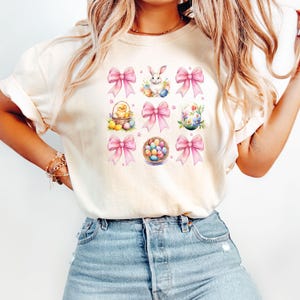 Puede incluir: Una camiseta blanca con un dise&ntilde;o de Pascua rosa y blanco. El dise&ntilde;o incluye un conejito, un pollito, una cesta de huevos y lazos rosas.