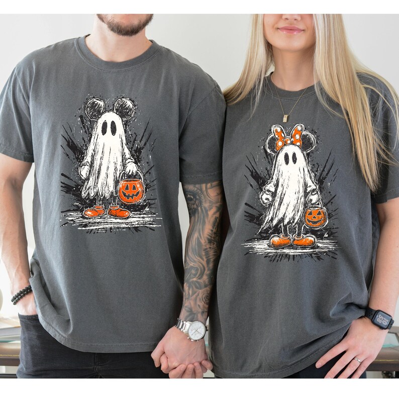May include: Two charcoal gray t-shirts featuring a cartoon ghost design with Mickey and Minnie Mouse ears. The ghosts are holding jack-o'-lantern buckets and wearing orange shoes. The design has a distressed, splattered background, perfect for Halloween.