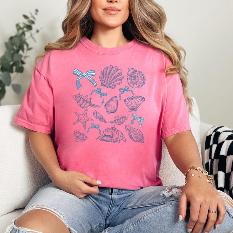 May include: A pink t-shirt with a blue and white seashell and bow design. The shirt is made of a soft, comfortable material and has a relaxed fit.