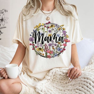 Peut inclure: Un t-shirt cr&egrave;me avec un motif de couronne de fleurs et le mot "Mama" au centre. Les fleurs sont dans des tons de rose, violet, jaune et bleu.
