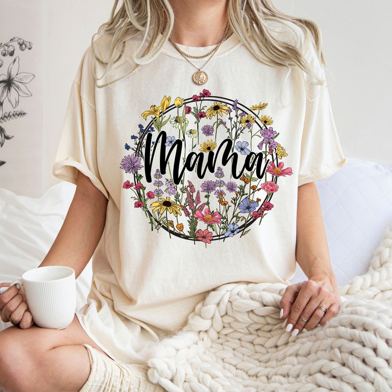 Peut inclure: Un t-shirt cr&egrave;me avec un motif de couronne de fleurs et le mot "Mama" au centre. Les fleurs sont dans des tons de rose, violet, jaune et bleu.