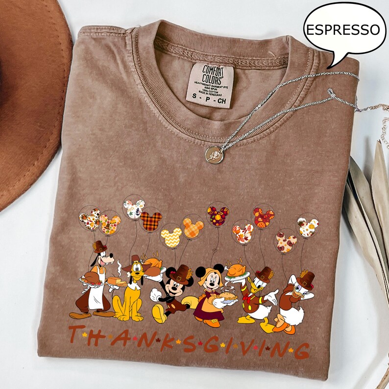 May include: Espresso-colored t-shirt featuring a Thanksgiving-themed graphic with Disney characters. The design includes Mickey Mouse, Minnie Mouse, Donald Duck, and Goofy. The word "THANKSGIVING" is printed across the bottom in brown letters. A silver necklace is draped across the shirt.