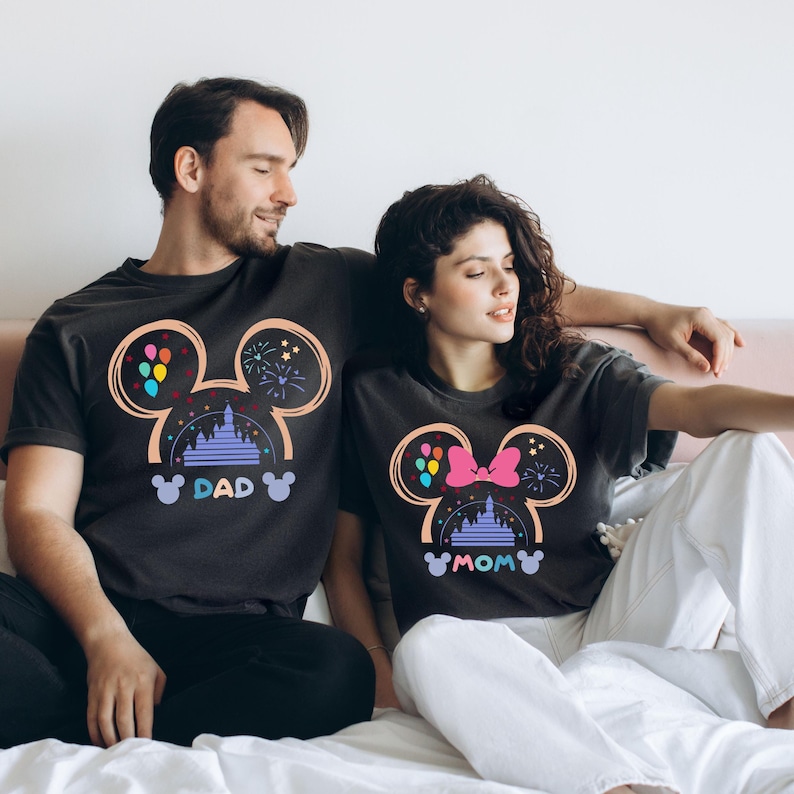 Pode incluir: Um casal vestindo camisetas pretas combinando com um design de silhueta do Mickey Mouse. As camisetas apresentam uma imagem de desenho animado do castelo da Cinderela, bal&otilde;es e fogos de artif&iacute;cio. A camiseta do homem diz "DAD" e a camiseta da mulher diz "MOM".