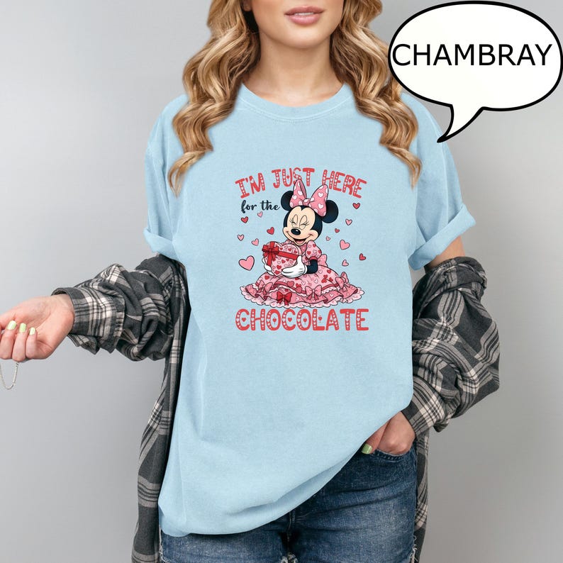 Puede incluir: Camiseta azul claro con un gr&aacute;fico de Minnie Mouse sosteniendo un regalo y el texto "I'm just here for the chocolate". El dise&ntilde;o presenta corazones rosas y un vestido rosa. La camiseta es de material suave.