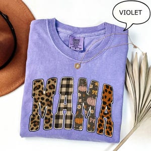 May include: A violet Comfort Colors t-shirt with the word "MAMA" in a mix of leopard print, plaid, and floral patterns. A gold necklace with a pendant rests on the shirt. A speech bubble above the shirt reads "VIOLET".