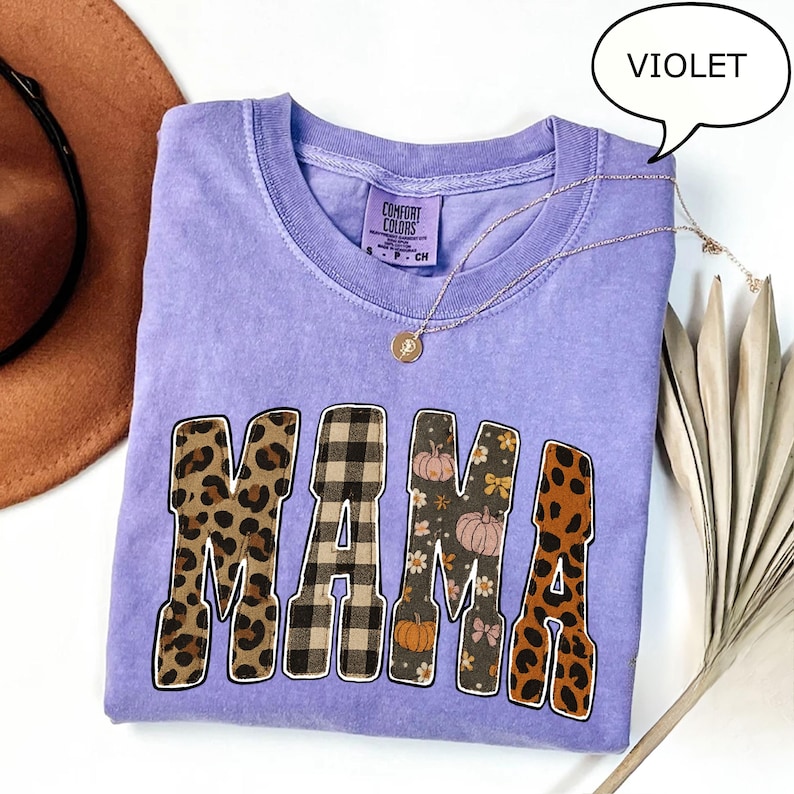 May include: A violet Comfort Colors t-shirt with the word "MAMA" in a mix of leopard print, plaid, and floral patterns. A gold necklace with a pendant rests on the shirt. A speech bubble above the shirt reads "VIOLET".