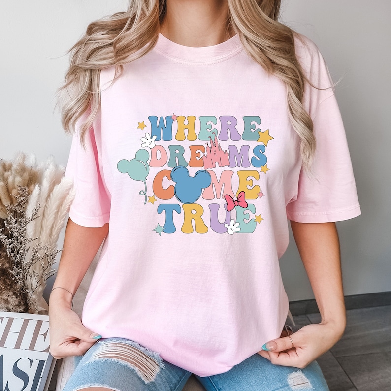 May include: Pink t-shirt with a graphic design that says "Where Dreams Come True" in colorful letters with a Mickey Mouse head and other Disney-themed icons.