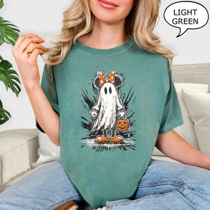 May include: A light green t-shirt featuring a cartoon ghost with Minnie Mouse ears and a bow. The ghost is holding a jack-o'-lantern bucket. The shirt has the text "LIGHT GREEN" in a speech bubble.